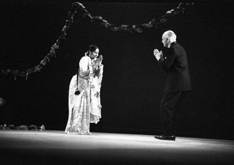 Black and white photo of T. Balasaraswati, Indian Bharatanatyam dancer, and Ted Shawn onstage in pools of bright light against a dark background with garland sweeping across behind them. Balasaraswati, at left, stands with her arms wreathed in a garland, bowing towards Shawn with her hands together in a prayer-type position. She is wearing her lusciously pleated white garments, having just finished performing. Shawn stands at the right side of the image in a tuxedo facing Balasaraswati and bows reverently at her mirroring her posture.
